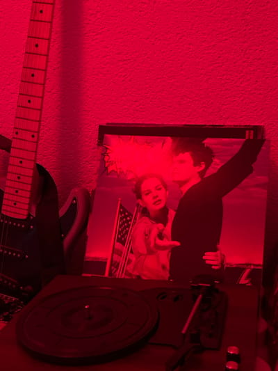 The scene, located in Kernen im Remstal, Germany, is characterized by a strong, uniform red illumination, casting a deep crimson hue over all visible objects against a textured wall. On the left, the neck and partial body of an electric guitar are visible, displaying a light-colored fretboard with dark dot inlays. Centrally positioned, a vinyl record album cover is propped upright. The album art features two figures: a woman on the left, seen from the waist up, extending her right arm forward with an open hand; and a man on the right, partially obscured, with his left arm raised high and his right arm around the woman's torso. An American flag on a flagpole is visible in the background behind the woman, set against a horizon. Above the woman's head, a jagged-edged speech bubble graphic is discernible. In the foreground, occupying the lower third of the image, is a dark turntable. Its circular platter, complete with a central spindle, is visible but currently holds no record. The tonearm is resting in its cradle on the right side of the turntable's base, alongside visible control knobs.