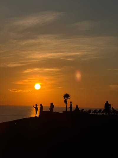 A vertical, medium shot captures a silhouette of a sunset over the sea, viewed from a high vantage point on Lý Sơn Island. The sun, a bright, glowing orb, hangs low in the sky, casting a warm, orange hue across the clouds and the ocean. Several figures, silhouetted against the vibrant sky, stand on the edge of a dark, elevated landmass, appearing to observe the sunset. Some are gathered in small groups, while others stand individually. A palm tree is also visible in silhouette, adding to the tropical feel of the scene. Rows of empty chairs are arranged on the elevated landmass, suggesting a viewing area set up for spectators. The sea stretches out to the horizon, meeting the sky in a soft gradient of colors. The overall impression is one of tranquility and natural beauty.