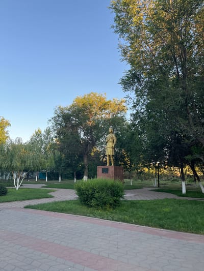 The image depicts a park setting in Qobda, Kazakhstan, bathed in the warm glow of late afternoon or early morning sun. A statue of a man in uniform stands prominently on a pedestal, surrounded by lush green grass and mature trees. A paved pathway, made of interlocking bricks with a reddish border, leads through the park, suggesting a well-maintained public space. The sky is a clear, bright blue, indicating fair weather. The overall scene conveys a sense of tranquility and historical commemoration. The statue is golden in color, with a plaque visible on its base. The surrounding vegetation is dense and varied, with some trees showing the warm hues of autumn foliage. A few white-painted tree trunks are visible, a common practice in some regions for protection or aesthetic reasons. The perspective suggests the photo was taken at ground level, looking up towards the trees and the statue. There are no discernible people actively engaged in an event, focusing instead on the static elements of the park.