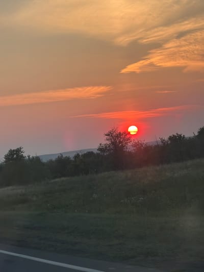 The image depicts a sunset over a rural landscape. The sun, a bright red orb with a yellow center, is positioned in the upper right quadrant of the frame, casting a warm glow across the sky. Wispy clouds, tinged with orange and pink, stretch across the sky. Below the sky, a dark treeline forms a silhouette against the horizon, with a hilly landscape visible behind it. In the foreground, a grassy field slopes downwards, with a white line, possibly a road marking, visible at the bottom left of the image. The overall scene is serene and natural, with no people or man-made structures other than the road marking. The image appears to be taken from a moving vehicle.