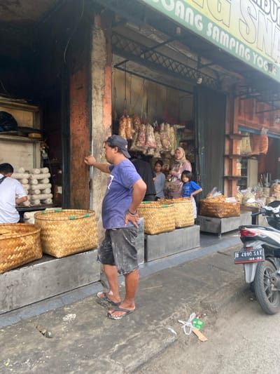 The scene is an outdoor market in Bojongsoang, Indonesia, located at coordinates -6.968 latitude and 107.637 longitude. There are five people standing outside a storefront. Three of the individuals appear to be customers. A man in a blue shirt is facing the storefront and engaging in a conversation with a woman in a head covering. Two children, one female and one male, are also in the conversation. A man in a grey shirt is standing behind the three of them. A man with a blue shirt and dark shorts is standing to the left and outside of the conversation. The storefront appears to be a food or produce market. Hanging from the ceiling, just inside the storefront, are strands of dried food, possibly noodles, beans, or spices. There are woven baskets stacked on the ground, and the sidewalk has a grey concrete surface. To the right of the group of people, a motorcycle is parked. The motorcycle has a license plate attached to the rear fender that reads "D 4990 SAT 03-25." The scene is lit by natural light, and it appears to be during the day.