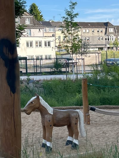 The image captures a wooden toy horse, predominantly brown with white and black accents, standing on a sandy surface in what appears to be a playground. The horse is the central subject, positioned slightly to the right of the frame. Behind the horse, a wooden post with a thick rope is visible, suggesting playground equipment. The background reveals a residential area with buildings, a fence, and lush green vegetation. A large tree is prominent on the left side of the image, partially obscuring the view of the buildings. The sky is blue with some clouds, indicating daylight. The overall scene suggests an outdoor recreational area in a suburban or urban setting, likely Waldbröl, Germany.