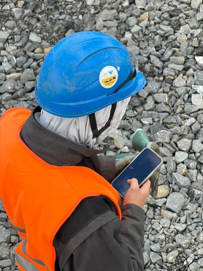 A person wearing a blue hard hat and an orange safety vest is looking at a smartphone. The person is also wearing gloves. The background is filled with gray rocks. The scene appears to be outdoors in an industrial or construction area. The person's face is obscured by a white cloth. The smartphone screen displays the words "Welcome back". A sticker is visible on the blue hard hat, which appears to be a company logo with text. The location is identified as Wilkowisko, Poland.