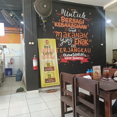 The image shows a restaurant interior in Kaliwungu, Indonesia, located at coordinates -6.962, 110.256. The restaurant is decorated with a black chalkboard on which is written a message in Indonesian which translates as: "It's time to share happiness through delicious and affordable food, by all Indonesian people." There is a red fire extinguisher next to a poster advertising a discount on food. Two wooden chairs sit in front of a wooden table that is partially visible. A person's hand holding a white plate is seen at the right edge of the image. In the background, a person can be seen sitting at a table to the right. Another person is visible in the far background. The floor is tiled in white.