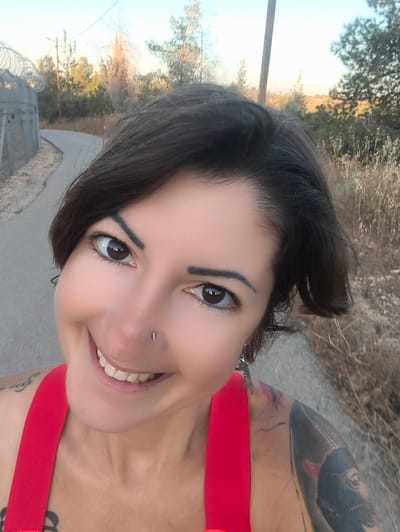 A medium shot photograph shows a woman with short, dark hair and brown eyes smiling at the camera. She is wearing a red top with thin straps, and a silver stud is visible in her nose. A large, dark tattoo covers her right arm and shoulder, and another smaller tattoo is visible on her left shoulder. She is outdoors, with a paved path and a wire fence visible behind her. The path curves to the left and appears to lead into a wooded area with dry grass and trees. The sky is a pale blue, suggesting either early morning or late afternoon light. The image appears to have been taken in Lachish Regional Council, Israel, based on the presence of a wire fence and arid vegetation characteristic of the region.