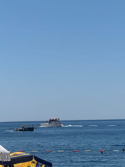 The image depicts a bright, clear day at a beach in Kamenovo, Montenegro. The sea is a deep blue, with gentle waves. In the distance, two boats are visible on the water. The larger boat, a white yacht, has a crowded deck with many people visible. It is moving to the right, leaving a white wake. The second, smaller boat is dark green and appears to be a fishing boat, also moving to the right with a few people on board. In the foreground, beach umbrellas are partially visible, one with a yellow and blue pattern, suggesting a recreational area. A rope with white and red buoys marks a boundary in the water, with a few people swimming or wading near it. The sky is a cloudless, pale blue.