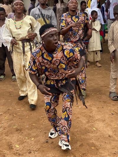 A group of people, some in traditional attire, stand on a dirt ground in Rigachikun, Nigeria. A young man in the foreground, wearing a colorful patterned outfit and a beaded headband, appears to be dancing or performing, with his mouth open and hands outstretched. Behind him, a woman in a golden traditional dress and beaded necklace stands with her eyes closed. Other individuals, including children and adults, are visible in the background, some wearing more modern clothing like a grey hoodie and others in white or beige garments. The environment is outdoors, with dry, dusty ground and scattered leaves.