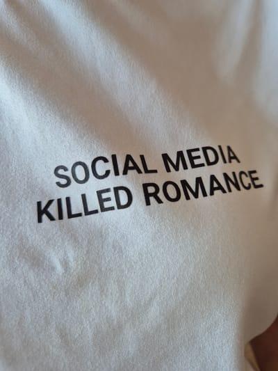 A close-up, low-angle shot shows the words "SOCIAL MEDIA KILLED ROMANCE" printed in black, sans-serif capital letters on a white fabric, likely a t-shirt. The fabric is creased and textured, with visible stitching lines. The lighting creates shadows and highlights, emphasizing the folds and contours of the material. There is a faint, light brown stain on the fabric below the text. The scene appears to be indoors, with no discernible external environmental features. The image does not depict any specific location in Voluyak, Bulgaria, or any individuals or actions.
