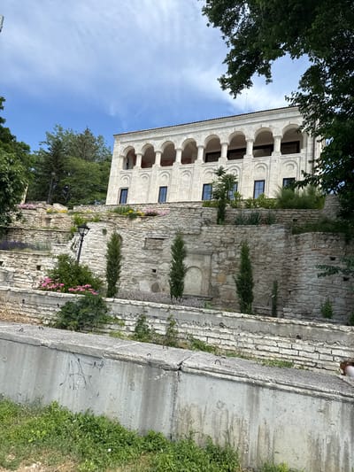 The image depicts a multi-level stone structure with arched windows and columns, possibly part of a historical building or ruins. The building is made of light-colored stone with arched openings and decorative elements above the windows. Adjacent to the main structure is a rough stone wall, overgrown with vegetation, including small trees and flowering plants. There are several lampposts visible, suggesting the area is accessible. The scene is set outdoors, with a bright blue sky and green trees surrounding the buildings. The presence of greenery and the clear sky suggest it is daytime with fair weather. In the foreground, a concrete wall with some graffiti is visible, along with grass and more vegetation. The overall impression is one of an old, weathered historical site being reclaimed by nature. The location is identified as Balchik, Bulgaria. No people are visible in the image, and no specific activity or event is taking place. The lighting and shadows indicate it is likely a sunny day.