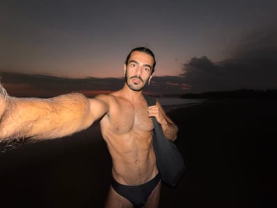 A man is captured in a selfie at a beach in Selemadeg, Indonesia, during dusk or twilight. He is bare-chested, sporting a beard and dark hair tied back, and wears a dark-colored speedo-style swimsuit. A dark fabric bag is slung over his right shoulder, and a small cross earring is visible in his left ear. His left arm is extended towards the camera, suggesting he is holding the device. The background features a twilight sky with a gradient of dark grey-blue transitioning to muted orange and pink hues near the horizon, with dark clouds scattered across the scene, particularly prominent on the right. A calm body of water is visible beyond the man, and a dark, indistinct landmass or tree line forms the distant horizon. The foreground, consistent with a beach, is dark. The man is directly illuminated, contrasting with the dim natural light of the environment.