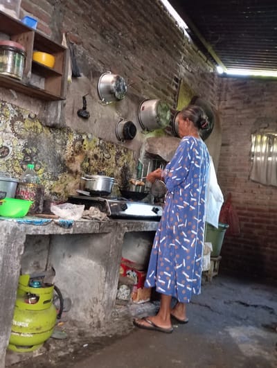 This image captures a domestic scene in what appears to be a humble kitchen or cooking area in Baron, Indonesia, during the daytime. The area is illuminated by bright natural light entering from an open doorway or window on the left, which features corrugated metal siding.
The primary subject is an older woman with gray hair tied back, wearing a blue and white patterned long dress (daster) and black flip-flops. She is actively engaged in cooking or food preparation, bending over a two-burner gas stove situated in the center-right of the frame. An aluminum pot is on the stove, and she seems to be attending to its contents.
The setting is characterized by its rustic and utilitarian nature. The floor is made of rough brick or concrete, partially covered by a stained, patterned mat around the cooking area. The walls are also constructed from exposed brick and rough concrete. Surrounding the woman and the stove are various household objects, including several aluminum pots and pans placed on the floor. To the right, a bright green propane gas cylinder is visible, connected to the stove, with the words "YA UNTUK" partially visible on its side.
The area exhibits a degree of clutter, with plastic bottles, plastic bags (one on the stove), and other miscellaneous items scattered about. In the lower-left foreground, a wooden shelf or divider holds a yellow bowl and another cylindrical container. Behind the woman, a white sheet or fabric and a reddish-brown sack are discernible. The overall impression is of a functional, albeit basic and somewhat untidy, cooking space within a modest dwelling.