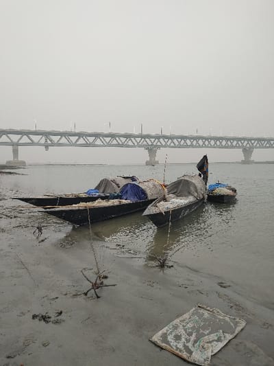 The image displays a riverine environment in Naodoba, Bangladesh, under an overcast, hazy sky. The foreground consists of wet, muddy earth. Several star-shaped mooring devices, appearing to be constructed from twisted metal or wood, are partially embedded in the mud, connected by ropes. A crumpled sheet of light-colored, textured material lies flat on the mud in the lower right. In the midground, three wooden boats are present: two larger, covered vessels are partially beached on the muddy bank, one featuring a visible blue tarpaulin and others covered in greyish material. A third, smaller boat floats in the calm, murky river water adjacent to the larger vessels. A single individual, dressed in dark attire, stands in the stern of the rightmost large boat, facing away from the viewer. The background features a prominent, multi-span steel truss bridge, supported by concrete piers, extending horizontally across the river. Numerous streetlights are visible along the bridge deck.