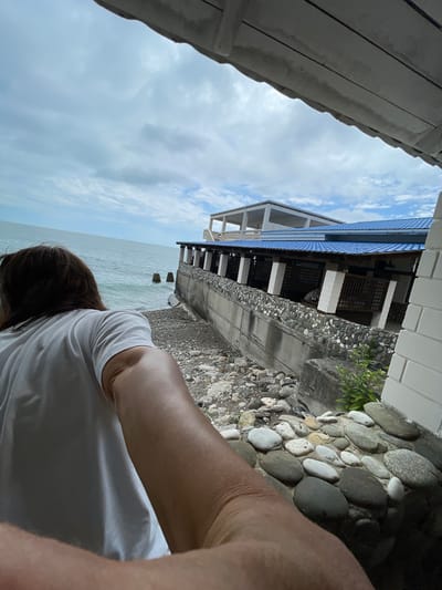 An over-the-shoulder, wide-angle shot captures a person in a white t-shirt looking out at the sea from a covered porch in Abaata, Abkhazia. The person's right arm and hand are extended towards the camera, obscuring the lower right corner. The sea, a tranquil blue-green, stretches to the horizon under a cloudy sky. Two weathered wooden posts stand in the water near the shore. A building with a blue tiled roof and open-air covered walkways is situated on a raised concrete embankment overlooking the sea. The embankment is lined with a wall constructed of grey concrete and large, smooth, multi-colored river stones. The ground leading down to the sea consists of similar stones and some gravel. The scene is set during the day, with overcast conditions.