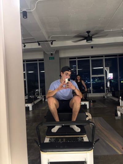 A man in a baseball cap and blue shirt poses for a selfie on a leg press machine at a gym. He wears white socks. In the background, another person works out near large windows showing a nighttime cityscape. The gym has high ceilings, exposed ductwork, and a ceiling fan. The image is a reflection in a mirror, with the bottom of the mirror visible in the foreground.