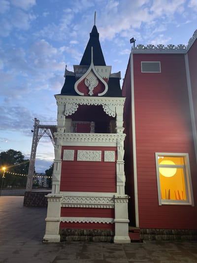 The image captures an outdoor scene in Vologda, Russia, at dusk. The primary subject is a traditional Russian wooden building with intricate white carvings and red siding. A prominent feature is a pointed roof, characteristic of older Russian architecture, possibly a small tower or part of a larger structure. A rectangular window on the upper part of the building emits a warm, yellow light, suggesting interior lighting is on.
Below the main building, a detailed, ornate wooden porch or balcony is visible, also adorned with white, carved embellishments and red wooden planks. The porch is supported by white pillars. The ground in front of the building is paved with rectangular stones.
In the background, through an opening in the ornate structure, a glimpse of trees, buildings, and string lights can be seen, indicating the building is situated in a public area or park. The sky is a soft blue with scattered white clouds, typical of twilight. A metal structure, possibly a lamppost or part of an overhead wire system, is also visible. The overall mood is peaceful and slightly nostalgic, with the warm interior light contrasting with the fading daylight. No people are visible in the frame.
