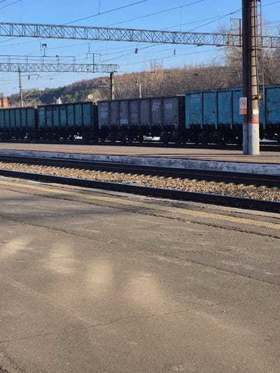 The image depicts a railway scene, likely in the city of Neftekamsk, Russia. A series of freight train cars, predominantly blue and gray, are stationary on the tracks. Parallel to the train tracks is a concrete platform marked with a yellow safety line. Overhead, a network of power lines and supporting pylons are visible against a blue sky. In the background, beyond the railway, there's a line of trees with bare branches. The general environment suggests an industrial or transportation area. No people are visible.