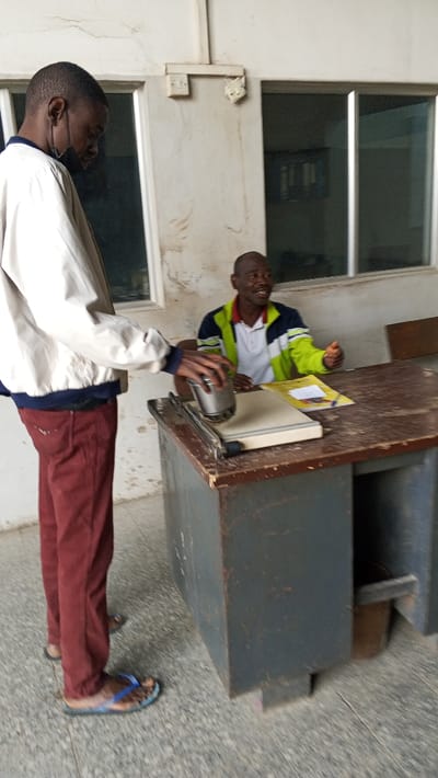 The image captures an indoor scene in Dakata, Nigeria, featuring two adult males interacting around a desk. A male subject stands on the left, clad in a light-colored jacket, maroon trousers, and blue-patterned flip-flops. He wears a black face mask lowered beneath his chin and is actively pressing a silver, cylindrical object onto a grey paper cutter or laminator situated on the desk. His attention is focused downward on this task. A second male subject is seated behind the desk to the right, wearing a white polo shirt layered with a green, blue, and white jacket. He is smiling and looking towards the standing individual, with his right hand resting on the dark brown, heavily worn wooden desk surface, near a yellow notebook and a pen. The desk itself has a dark grey base. The background consists of a light-colored wall exhibiting stains and cracks, with multiple rectangular windows featuring dark frames. A white electrical light switch and a damaged fixture are visible on the wall above the windows. The floor is tiled in grey. The overall environment suggests an operational, possibly public or administrative, space showing signs of age and use.