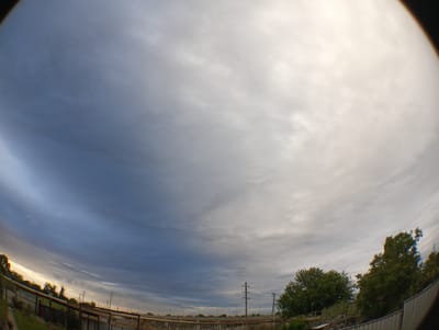 The image depicts a wide-angle view of the sky over a rural or semi-urban area, likely on the outskirts of Krasnyye Barrikady, Russia, given the context. The sky is filled with a dramatic display of clouds, with a gradient ranging from dark blue on the left to bright white and yellow on the right, suggesting either sunrise or sunset. The clouds are textured and appear to be in motion. In the lower portion of the image, a fence runs horizontally across the frame, made of metal poles and horizontal bars, with some yellow piping also visible. Beyond the fence, there are some trees and vegetation, appearing somewhat sparse. Several utility poles with wires are scattered throughout the scene, indicating the presence of electrical infrastructure. The overall impression is of an open, possibly industrial or agricultural landscape under a dynamic sky. There are no people or animals visible in the image, and no discernible actions or interactions are taking place. The environment appears to be outdoors, with a sense of natural light.