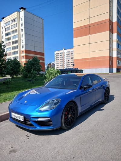 A bright blue Porsche Panamera is parked on asphalt pavement in front of a residential building in Kemerovo, Russia. The car has black rims and red brake calipers. The building behind the car is a multi-story structure with beige and orange colored sections and numerous windows. The sky is clear and blue. In the background, more residential buildings and green trees are visible. The license plate on the car is clearly visible and reads "C666YE 42" and "RUS". There are some dark spots on the pavement in front of the car that appear to be oil stains.
