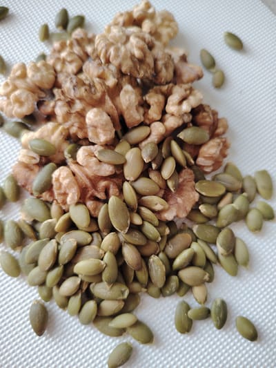 This is a close-up, top-down shot of a pile of walnuts and pumpkin seeds on a white textured surface. The walnuts are a light brown color and have a wrinkled, irregular shape, appearing to be shelled. The pumpkin seeds are a muted green color and are elongated and flat. They are scattered amongst and around the walnuts, with some appearing to be partially covered by the walnuts. The lighting is bright and even, suggesting it might be daytime with diffused light. The background is a white, woven-like material that provides a neutral backdrop, allowing the subjects to stand out. No people, text, or specific location cues are visible in the image. The overall impression is a simple arrangement of healthy food items.