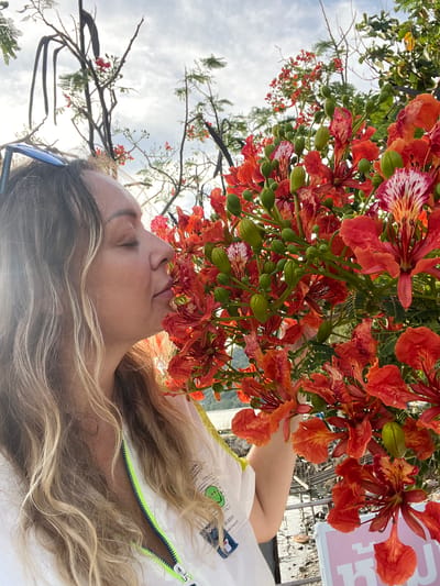 A woman with blonde hair is standing in front of a tree with bright orange flowers. She is closing her eyes and smelling the flowers. She is wearing a white jacket with a neon green zipper. The background is a cloudy sky with some green trees and a body of water. A sign with Thai writing is partially visible in the lower right corner of the image. The scene is located in Ban Haad Rin, Thailand.