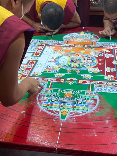 The image shows a group of people, likely Buddhist monks given their attire, working on a complex and colorful sand mandala. The mandala is constructed on a red-painted surface, and its design features intricate geometric patterns, floral motifs, and symbolic representations, predominantly in shades of green, white, red, blue, orange, and yellow. At least two monks are actively engaged in adding details to the mandala, one holding a tool, possibly a Chak-Pur (a metal funnel) used to deposit sand, and the other pointing, likely directing the placement or design of the sand. The environment suggests an indoor setting, possibly a monastery or a religious site. It can be reasonably inferred from the cultural context of sand mandalas that this activity is happening in Dharamshala, India, as it is a significant center of Tibetan Buddhism in exile.
