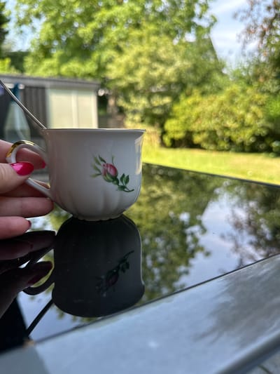A person's hand with bright pink fingernails holds a white teacup with a gold rim and a pink rose design. A silver spoon is placed in the cup. The cup is resting on a reflective black surface, creating a clear reflection of the cup and the rose design. The background is blurred and shows lush green trees and a light-colored structure. The scene appears to be outdoors on a sunny day. No specific Krefeld location is identifiable from the image.