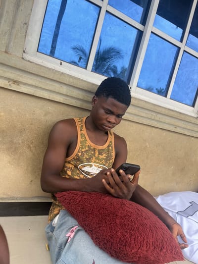 A young Black male, appearing to be in his late teens or early twenties, is seated in a semi-outdoor or sheltered area in Ikot Ekpene, Nigeria. He wears a camouflage-patterned tank top in olive green, brown, and tan hues, and light blue ripped jeans with a pink design on the left leg. A small gold stud earring is present in his left ear. He is actively engaged with a black smartphone, holding it with both hands and looking down at its screen with a focused expression. A dark red, fluffy pillow rests on his lap, and a white garment with a partial grey "1" visible is to his right. The background features a light brown, textured wall with horizontal wooden molding above it. Above the molding is a window with white frames and multiple panes of blue-tinted glass, reflecting palm tree fronds and possibly a vertical structure. The scene is illuminated by natural light.