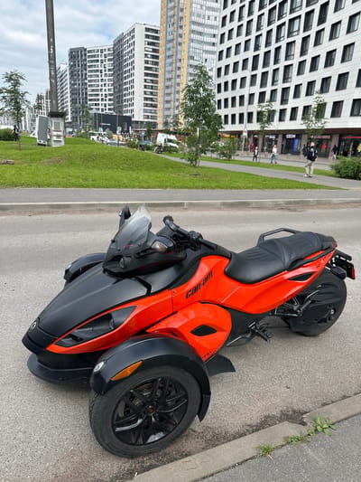 A red and black Can-Am Spyder three-wheeled motorcycle is parked on a paved road in front of a sidewalk and a grassy embankment. The motorcycle is positioned at an angle, facing slightly to the right. In the background, there are modern high-rise buildings, characteristic of the city of Murino, Russia. People are walking on the sidewalk, and some cars are visible on the road behind the motorcycle. The sky is overcast, and the overall lighting suggests it is daytime.