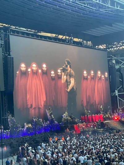 The image depicts an outdoor concert stage with a large video screen in the background. On the screen, a performer wearing a vest and dark pants, holding a microphone, stands in front of a group of figures in flowing red robes and gold masks. The stage below the screen features a set designed to look like ruins, with blue and red lighting illuminating the structures. A large crowd of concertgoers is visible in the foreground, facing the stage. The overall impression is of a dynamic live performance. The location is not specified in the image.