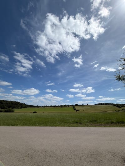 This image presents a wide-angle landscape scene captured in Niederanven, Luxembourg, on a clear day. The lower approximately one-fifth of the frame is occupied by a light grey asphalt or compacted gravel road, showing a granular texture and appearing empty. Above the road, expansive green fields stretch towards the distant horizon, exhibiting varied shades of green indicative of different agricultural sections or natural grasslands. Dark green forested areas and treelines are visible in the mid-ground and background, particularly a dense woodland strip on the far left. The sky, which comprises the upper half of the image, is a vivid blue with numerous scattered white clouds ranging from wispy cirrus to more defined cumulus formations. A prominent lens flare is observable in the upper right quadrant, suggesting the sun's position. No individuals, animals, or artificial structures are present in the visible landscape.