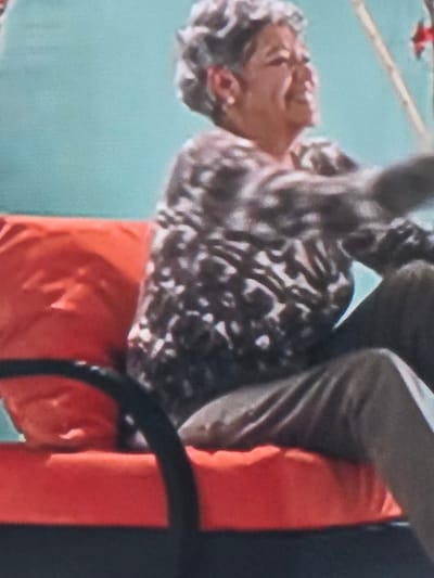 The image depicts an elderly woman seated outdoors against a light blue-green background, which has a visible fabric-like texture. The woman, shown from the chest up, has short, curly grey hair and wears a white stud earring in her visible left ear. She is smiling broadly and looking towards the upper right quadrant of the frame. Her attire consists of a long-sleeved shirt with a mottled dark brown and light grey or white pattern, and light-colored trousers. She is seated on a bright orange cushion, which appears to be part of a larger outdoor seating unit, possibly a swing or bench, given the black curved armrest visible in the lower foreground. In the upper right corner, a thin, light-colored vertical pole or stick is partially visible, with a small, indistinct red object attached to it. The environment is brightly lit, suggesting daylight. The image does not contain visual cues to confirm its specific location within the city of Kyustendil, Bulgaria, as requested.