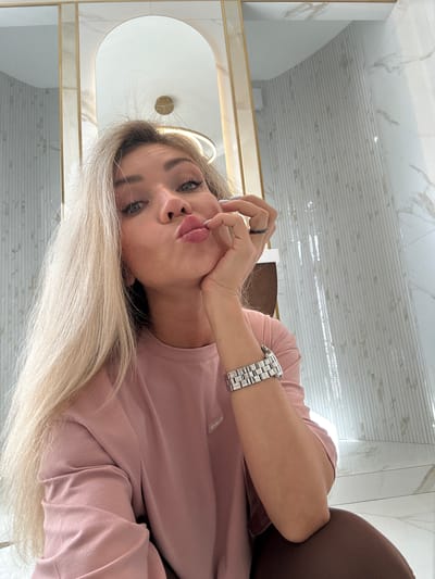 A blonde woman with fair skin and green eyes is taking a selfie in what appears to be a bathroom. She is wearing a pink long-sleeved t-shirt and a silver watch on her left wrist. Her right hand is resting on her chin, and she is puckering her lips. The background consists of marble-tiled walls and a large mirror with a golden frame. The lighting is bright, suggesting it is daytime. The location in the city of Armavir, Russia is not ascertainable from the image.