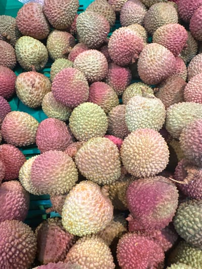 The image displays a close-up, overhead view of a large pile of fresh lychees. The lychees are a mix of colors, predominantly pinkish-red and pale green, with some appearing more yellow or even white. Their characteristic bumpy texture is clearly visible. The fruits are tightly packed, suggesting they are in a market stall or a retail display. The background appears to be a green plastic crate, further indicating a market setting. There are no people, actions, or interactions depicted in the image. The image does not contain any information about the city of Усмангали, Russia.