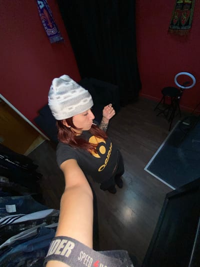 A low-angle, indoor shot depicting a person in a black t-shirt with gold lettering lying down. The person wears a gray and white beanie. Red hair is visible. The arm closest to the camera reveals an "UNDER" underwear band. The surrounding environment includes a red wall, a black blanket, wooden floor, a clothing rack with hanging clothes, a ring light and microphone stand. The location is likely a bedroom or studio in Llavallol, Argentina.