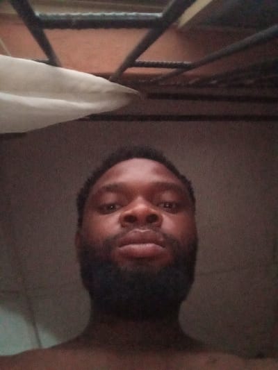An upward-angle close-up photograph depicts an adult male, likely of African descent, centered in the frame. He has dark skin, short dark hair, and a full, dark beard. His eyes are open and directed towards the camera, with a neutral expression. Only his head, neck, and upper chest are visible; no specific clothing is discernible. The background is dimly lit and out of focus. Above his head, a structure composed of dark, intersecting metal bars is visible against a lighter, possibly orange-brown, ceiling or surface. To the upper left, a portion of a white, folded or draped fabric object is present. The overall lighting is low, contributing to a grainy image quality. The scene appears to be an indoor setting in Magboro, Nigeria.