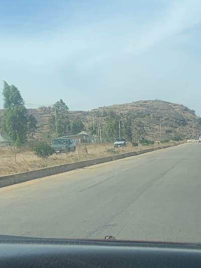 A landscape image shows a rocky hill beside a paved road. The road is gray and takes up a large portion of the right side of the frame. On the other side of the road is a steep, rocky hill with sparse vegetation. Several buildings and vehicles are situated at the base of the hill along the roadside. Power lines stretch across the scene. The sky is pale and clear. The photo is taken from inside a vehicle, with a portion of the car visible on the right edge.