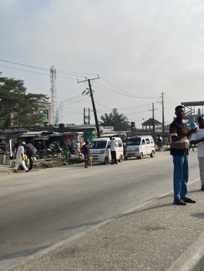 A bustling street scene unfolds with a row of white vans parked along the roadside. Several people are visible, including two men near the vans and another walking along the pavement with a blue water bottle. Adjacent to the road is a lively commercial area with stalls and various vehicles. Overhead, a network of power lines and a communications tower are present, contributing to the urban landscape under a partly cloudy sky.