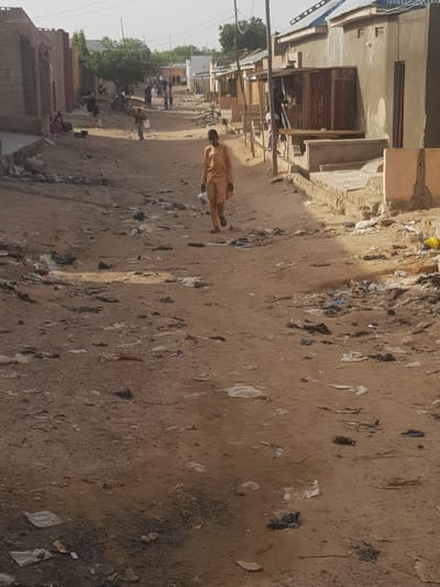 The image depicts a street scene in Dabawa, Nigeria. The unpaved road is littered with garbage and debris. A man in an ochre-colored garment walks down the center of the street. Other people are visible further down the street near some trees, appearing as blurred figures in the distance. Buildings of modest construction line both sides of the street, some with corrugated metal elements. The overall environment suggests a low-income or impoverished neighborhood.
