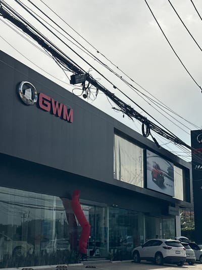 The exterior of a GWM car dealership is visible, with the GWM logo and red lettering prominently displayed on the dark grey facade. A large digital billboard on the building shows an advertisement for a red GWM TANK 300. The building features large glass windows, reflecting the surroundings, and grass is planted along the base. A bright red inflatable tube dancer is positioned in front of the glass, its form bent and distorted. Several cars are parked outside the dealership, including a white SUV. Above the building, a dense network of electrical wires crisscrosses against a bright, overcast sky. The overall scene suggests a commercial establishment in an urban or suburban setting, likely in Kathu, Thailand, given the context.