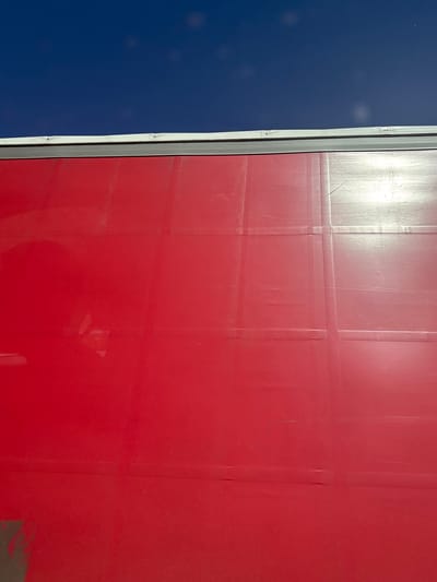 The image shows an exterior view of a commercial building. The exterior is red paneling. The paneling features a grid pattern. Next to the red paneling, there is a narrow grey metal trim. To the left of the trim, the building exterior is a solid blue color. There is a bright spot in the top center of the image due to the sunlight. The specific location of the scene in the city of Meyreuil, France, cannot be determined from the image.