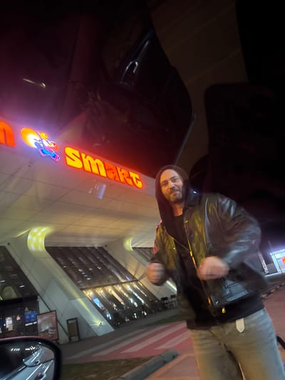 A man wearing a black leather jacket and gray jeans stands near a building with a large illuminated sign that says "Smart" vertically. He is smiling and facing the camera. The building has numerous windows. A car's side mirror is visible in the lower right corner of the frame. The overall setting appears to be at night.