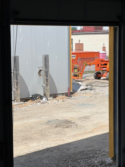 The image depicts an outdoor construction site viewed from inside a building. In the foreground, a light gray corrugated metal wall is visible, with two tall, reflective metal pipes extending vertically from its base. The ground in front of the wall is covered with gravel and construction debris.
Beyond the wall, an orange shipping container labeled "Boels RENTAL" is present, along with an orange boom lift. In the background, a light-colored building with red-tiled roofing and several windows and doors can be seen. The scene is well-lit by sunlight, casting shadows across the ground.
The location is identified as Modřice, Czechia. The overall impression is one of ongoing construction work in an industrial or commercial area. There are no people visible in the image.