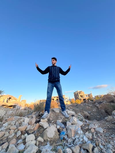A male individual is depicted standing on a mound of light-colored rubble in Joubar Municipality, Syria. The subject, with dark hair and a light beard, wears a dark blue sweater with light geometric patterns, blue jeans, and grey athletic shoes. He stands with his legs slightly apart, both arms bent at the elbows, and his index fingers pointing upwards. His head is turned slightly to his right, looking off-frame. The background features an extensive urban landscape of severely damaged and ruined multi-story buildings, characterized by missing walls, exposed frameworks, and scattered debris. Sparse dry vegetation is present throughout the scene. The lighting suggests afternoon conditions under a clear, light blue sky. Visible detritus in the foreground rubble includes plastic bottles and other discarded materials.