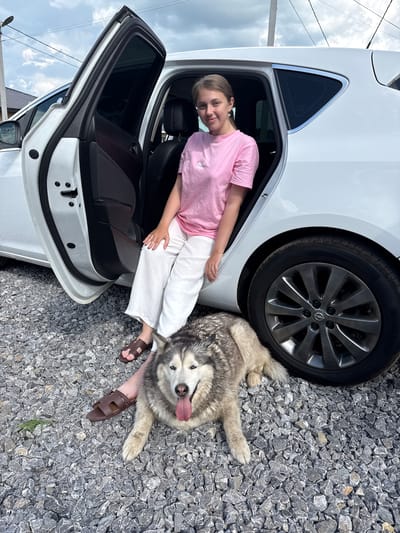 In Нагаево, Russia, on a bright, overcast day, a young woman with light brown hair, wearing a pink t-shirt and white pants, sits on the open driver's side door of a white car. She is positioned with her legs extended outward, one foot resting on the car's frame and the other on the ground. Beside her, on the gravelly ground, lies a husky dog, its tongue hanging out. The car is parked in an outdoor area, indicated by the gravel and the faint outline of a building and telephone pole in the background. The dog appears relaxed, lying on its side with its paws extended, while the woman looks towards the camera with a slight smile. The scene captures a casual moment of companionship between the woman and her dog in front of a parked vehicle.