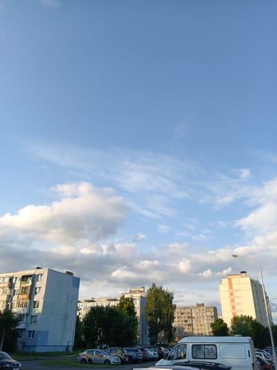 The image depicts a residential area in the city of Baranavichy, Belarus, under a partially cloudy blue sky. In the foreground, a paved area shows several parked cars, including a white van, a silver sedan with taxi markings, and other vehicles. To the left, a multi-story apartment building with a light blue and gray facade is visible. Behind it, another apartment building of similar height with a gray facade is partially obscured by trees. Further to the right, a taller, cream-colored apartment building with orange vertical stripes stands out. The environment appears to be a typical urban setting with residential blocks and a parking lot. The time of day appears to be late afternoon or early evening, judging by the lighting.