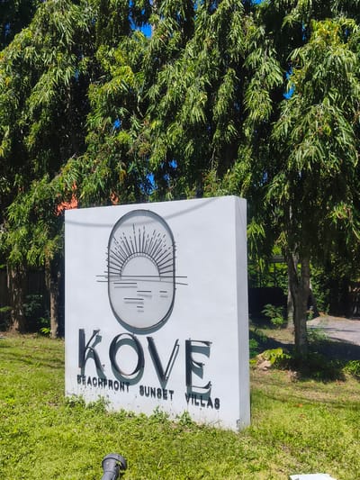 In the city of Baan Thong Po, Thailand, a white rectangular sign stands on a grassy area, partially obscured by lush green foliage from overhead trees. The sign displays the name "KOVE" in large, black, metallic letters. Below "KOVE," in smaller black text, are the words "BEACHFRONT SUNSET VILLAS." To the left of the text, the sign features a circular emblem containing a stylized depiction of a setting sun over water, rendered with thin, dark lines. The environment is sunny and appears to be an outdoor setting, likely a public space given the sign.