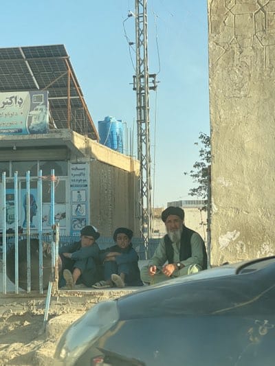 In Arghandab, Afghanistan, three males sit on a curb along a street. Two younger males are on the left, wearing dark-colored clothing and hats. An older male with a white beard and a black turban sits on the right, wearing a light-green vest over a dark-colored garment. In the background is a building with a sign in a non-English language, topped with a solar panel array. A utility pole stands tall near the building, and a blue water tank is visible on the roof. Part of a vehicle is visible in the foreground. The sky is clear and blue.