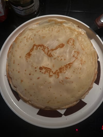 The image shows a stack of crepes on a white plate with a black and white geometric pattern on the rim. The crepes have a light brown color and a slightly uneven texture. There is a dark object, possibly a cooking appliance, in the upper right corner of the image. To the lower left, there's a jar labeled "TERRA DI SANREMO" and part of another package.

Without further information or context, it is impossible to confirm the location is Sanremo, Italy or to identify the actions, interactions, or people present in the scene beyond the crepes and the surrounding objects.