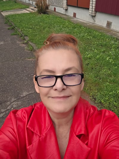A woman with auburn hair pulled into a bun and wearing glasses and a red leather jacket stands outside. The background shows a sidewalk, grass, and a building. The location is Daugavpils, Latvia.